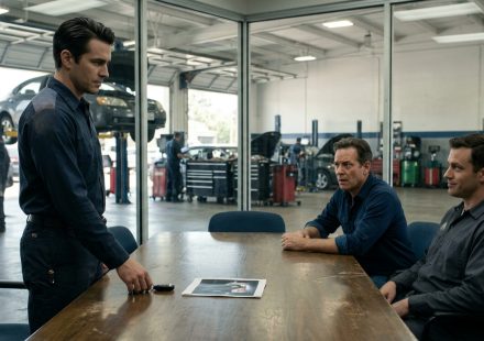An automotive garage manager calmly faces dismissal as the owner points to a photograph accusing the employee of working weekends at another garage.