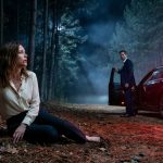 A frightened woman sits beside a secluded forest road at night while her husband stands near a luxury sedan after abandoning her during a medical emergency.
