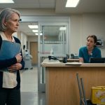 A worried grandmother holding a blue medical folder stands outside the glass doors of a hospital intensive care unit while a nurse explains a visitor restriction.