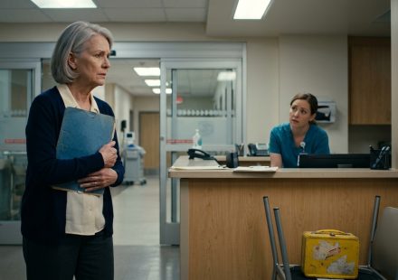 A worried grandmother holding a blue medical folder stands outside the glass doors of a hospital intensive care unit while a nurse explains a visitor restriction.