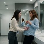 A frightened pregnant office worker pulls away as a female coworker places her hands on her stomach inside a modern corporate bathroom.