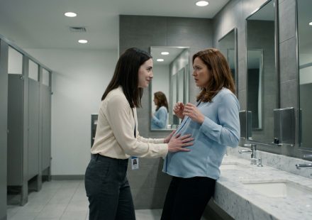 A frightened pregnant office worker pulls away as a female coworker places her hands on her stomach inside a modern corporate bathroom.
