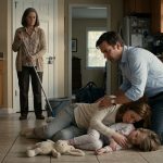 A shocked father kneels beside his weak wife and six-year-old daughter on their kitchen floor while his mother stands nearby holding a mop.