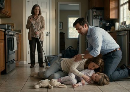 A shocked father kneels beside his weak wife and six-year-old daughter on their kitchen floor while his mother stands nearby holding a mop.