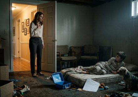 A concerned aunt kneels beside her seriously ill nephew in a neglected room, with a blue lunchbox and suspicious school withdrawal paperwork nearby.