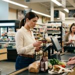 A 31-year-old woman looks concerned while holding a declined debit card and smartphone at a grocery store checkout counter, with groceries waiting beside the register.