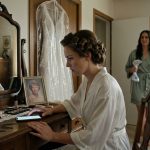 A bride in a white robe sits at her vanity staring suspiciously at an illuminated smartphone while her maid of honour returns to the bedroom.
