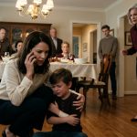 A mother kneels beside her injured seven-year-old son in a family dining room while her angry mother confronts her for calling emergency services.
