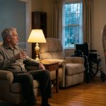 A shocked elderly husband watches his wife stand and walk across their living room while her empty wheelchair sits beside a recliner.
