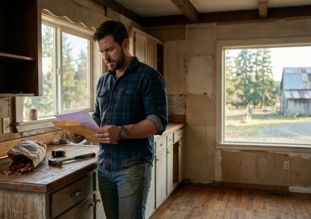 A hardworking man stands inside his inherited rural farmhouse, holding legal documents beside a kitchen counter with tools and a handwritten note, looking stunned by his family's demand.