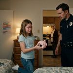 A concerned father in a police uniform comforts his twelve-year-old daughter as she holds an empty pink piggy bank in her bedroom, while her grandmother stands in the kitchen.