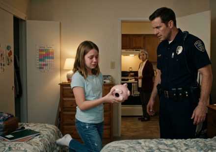 A concerned father in a police uniform comforts his twelve-year-old daughter as she holds an empty pink piggy bank in her bedroom, while her grandmother stands in the kitchen.