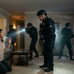A frightened mother kneels in her living room as armed SWAT officers enter her home at night while a police sergeant approaches her.