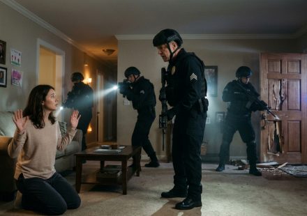 A frightened mother kneels in her living room as armed SWAT officers enter her home at night while a police sergeant approaches her.