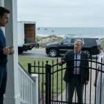 A man holding a coffee mug confronts his wealthy father and older sister outside a luxury beach house, with two moving trucks parked at the gate.