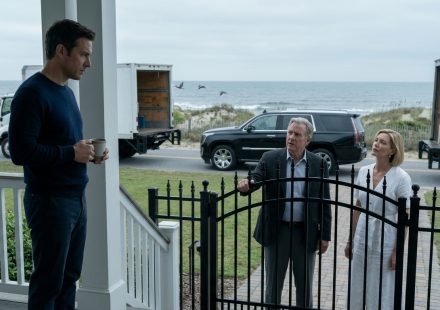 A man holding a coffee mug confronts his wealthy father and older sister outside a luxury beach house, with two moving trucks parked at the gate.