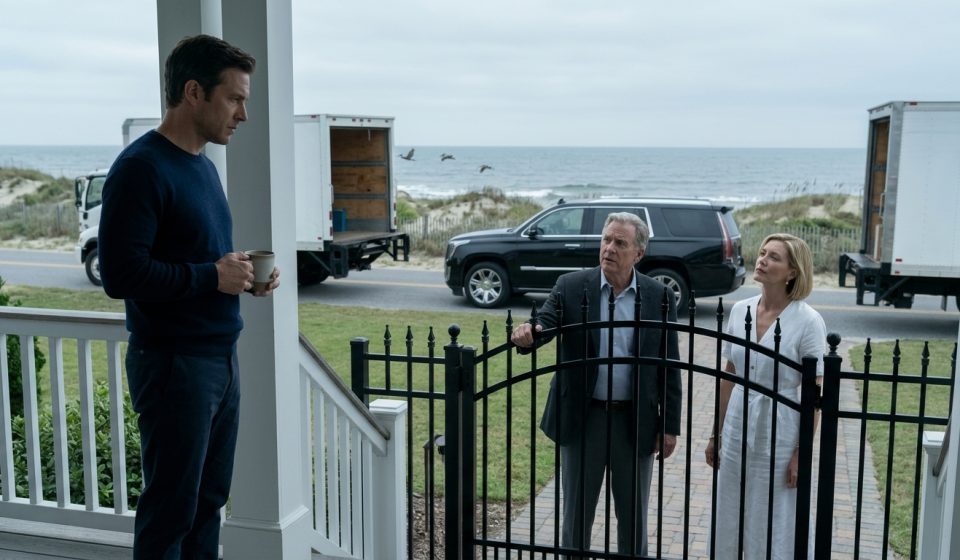 A man holding a coffee mug confronts his wealthy father and older sister outside a luxury beach house, with two moving trucks parked at the gate.