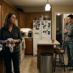 A stepmother stands in her kitchen holding a dish towel while her teenage stepson in a grey hoodie faces her with his arms crossed.