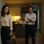 A frightened professional woman standing in her dimly lit apartment doorway while her angry husband confronts her about arriving home late from work.