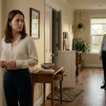 A concerned 32-year-old woman stands inside her suburban home, noticing a slightly open door and misplaced belongings while her husband remains in the background.