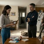 A shocked 21-year-old woman opens a birthday gift box containing a one-way bus ticket to Denver while her father stands with crossed arms and her sister smirks.