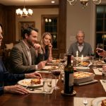 A composed woman sits at an elegant dinner table while her husband and his friends laugh, as an older guest in a gray cardigan quietly observes.