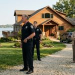 A shocked homeowner stands outside her lakefront vacation house facing two police officers, while her mother and sister watch from the driveway during a family celebration.