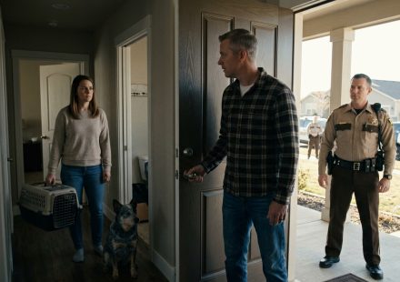 A woman standing beside her dog's crate in a hallway as her husband opens the front door to a sheriff's deputy, with patrol cars parked outside.