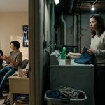 A stepmother stands beside a clothes dryer holding jeans, shocked by a conversation coming through a forgotten baby monitor in her basement laundry room.