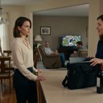 Two adult sisters stand tensely in a family kitchen beside a portable computer monitor and laptop bag as a promotion celebration turns uncomfortable.