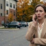 A twenty-seven-year-old professional woman standing alone in an autumn apartment parking lot in Vermont, staring at her phone after a painful conversation with her mother.
