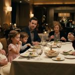 An elderly father looks shocked as his demanding son-in-law confronts him across a dining table while his daughter sits silently in their family home.