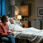 An exhausted daughter sits beside her elderly father's hospice bed as her wealthy brother stands nearby in a charcoal suit, smiling inappropriately.